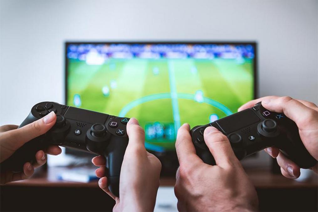 closeup of two sets of hands holding gaming controls with a TV and game on in the background