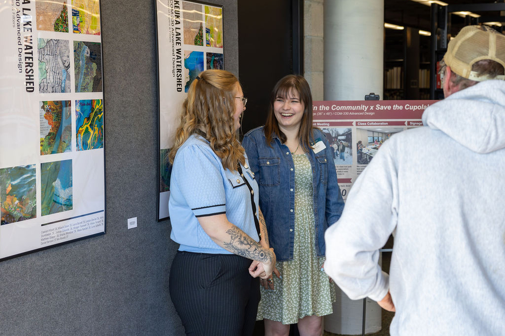 Two people stand smiling and talking beside framed posters and a presentation board at an indoor academic showcase or exhibition. A third person, seen from behind, appears to be engaging with them. The display features environmental and watershed-related projects, creating a welcoming and collaborative atmosphere.