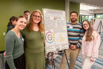 Four college students stand smiling on either side of a research poster displayed on an easel in a campus hallway. The poster discusses occupational therapy, student belonging, and wellness, with charts and text sections. The students wear name tags and casual attire, and other attendees are visible in the background of the indoor event space.