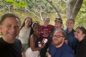 A group of students and two adults pose for a selfie beside a calm river surrounded by dense tropical trees and branches. Everyone is smiling, with some wearing glasses or sunglasses, and the group stands closely together on a shaded wooden overlook.
